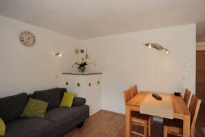A room with a grey sofa, a wooden dining table, a wall clock, and a small shelf with decorative items.