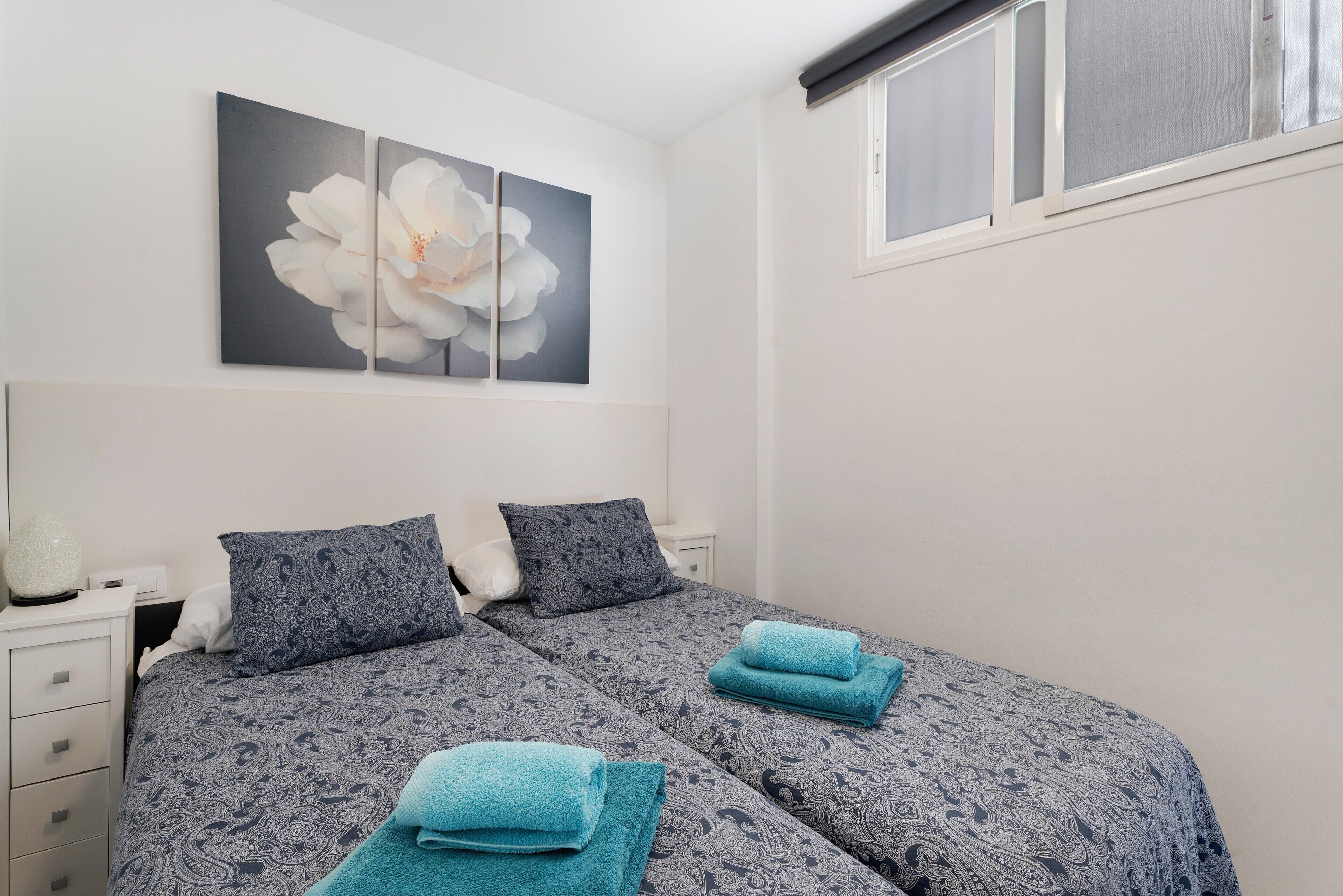 A bedroom with two beds, a nightstand, a lamp, and a large floral artwork on the wall.