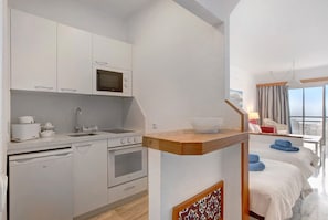 Une cuisine moderne avec des meubles blancs, un four à micro-ondes et un évier. On y trouve une étagère en bois sur laquelle est posé un bol. La pièce comprend un lit avec une literie blanche et une fenêtre offrant une vue sur le ciel.