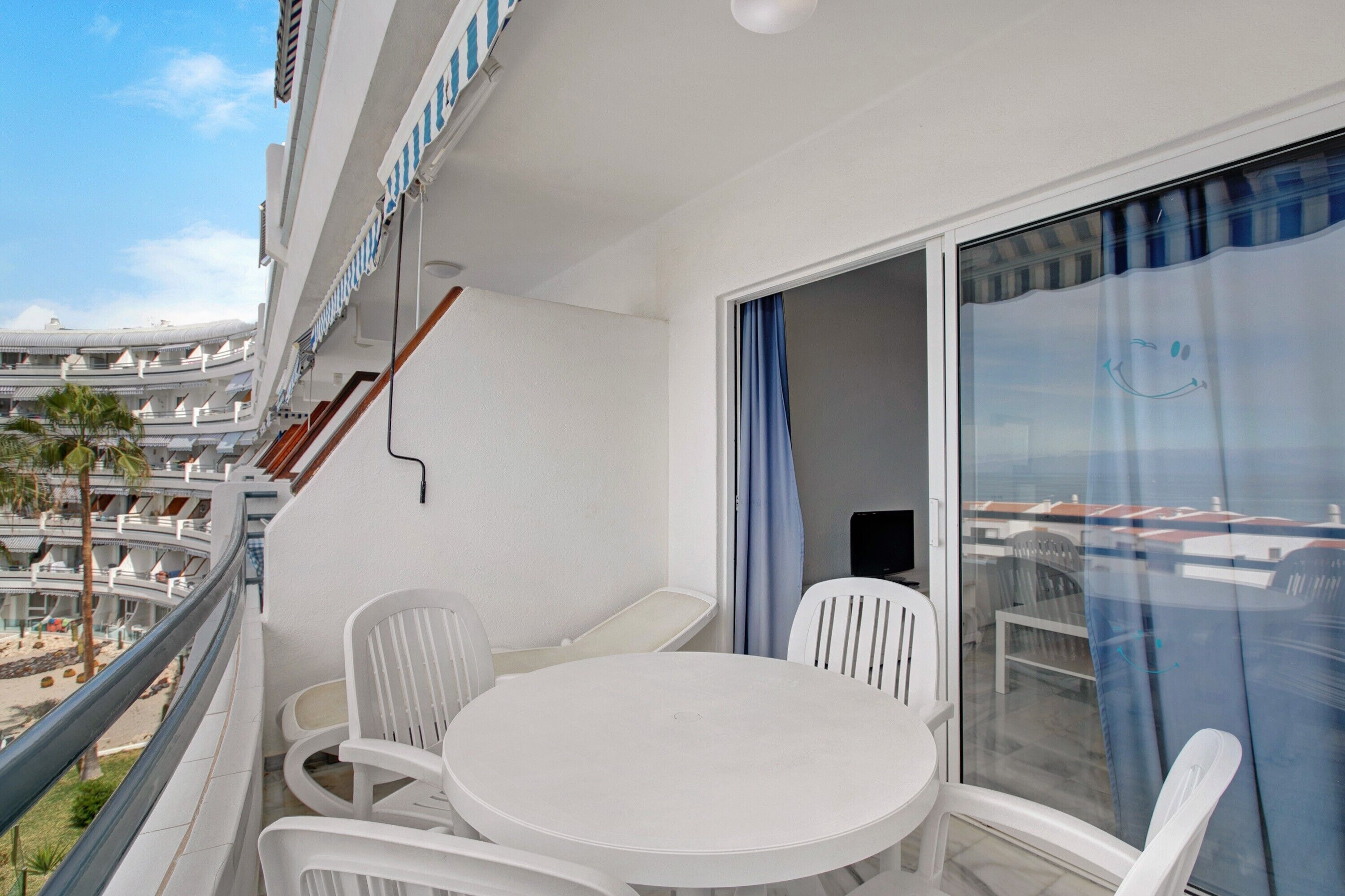 Un balcon doté d’une table ronde et de chaises blanches, offrant une vue sur une marina avec des bateaux et un immeuble à multiples balcons.