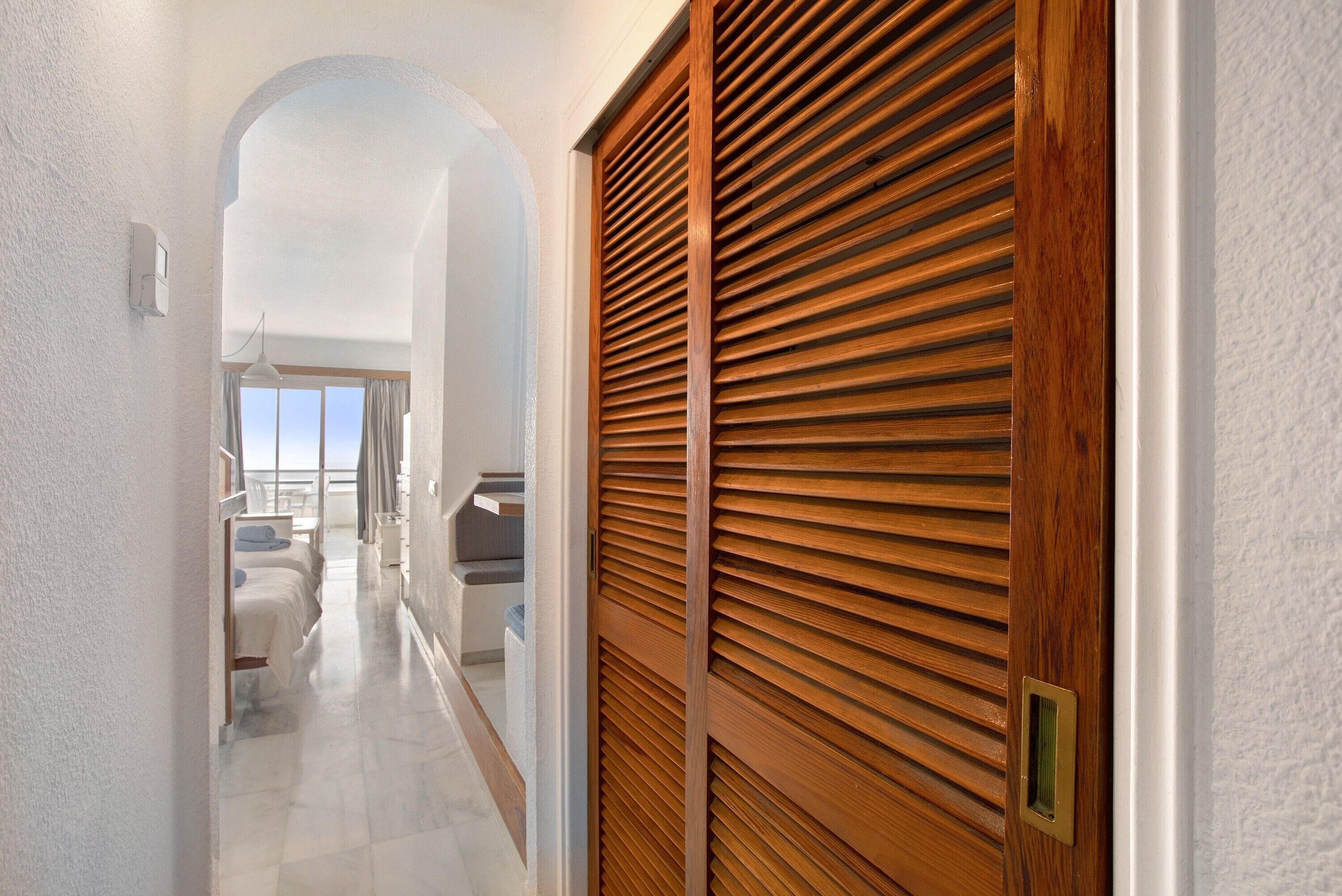 Un couloir avec une porte en bois, un mur blanc et une vue sur un balcon doté d’une balustrade.