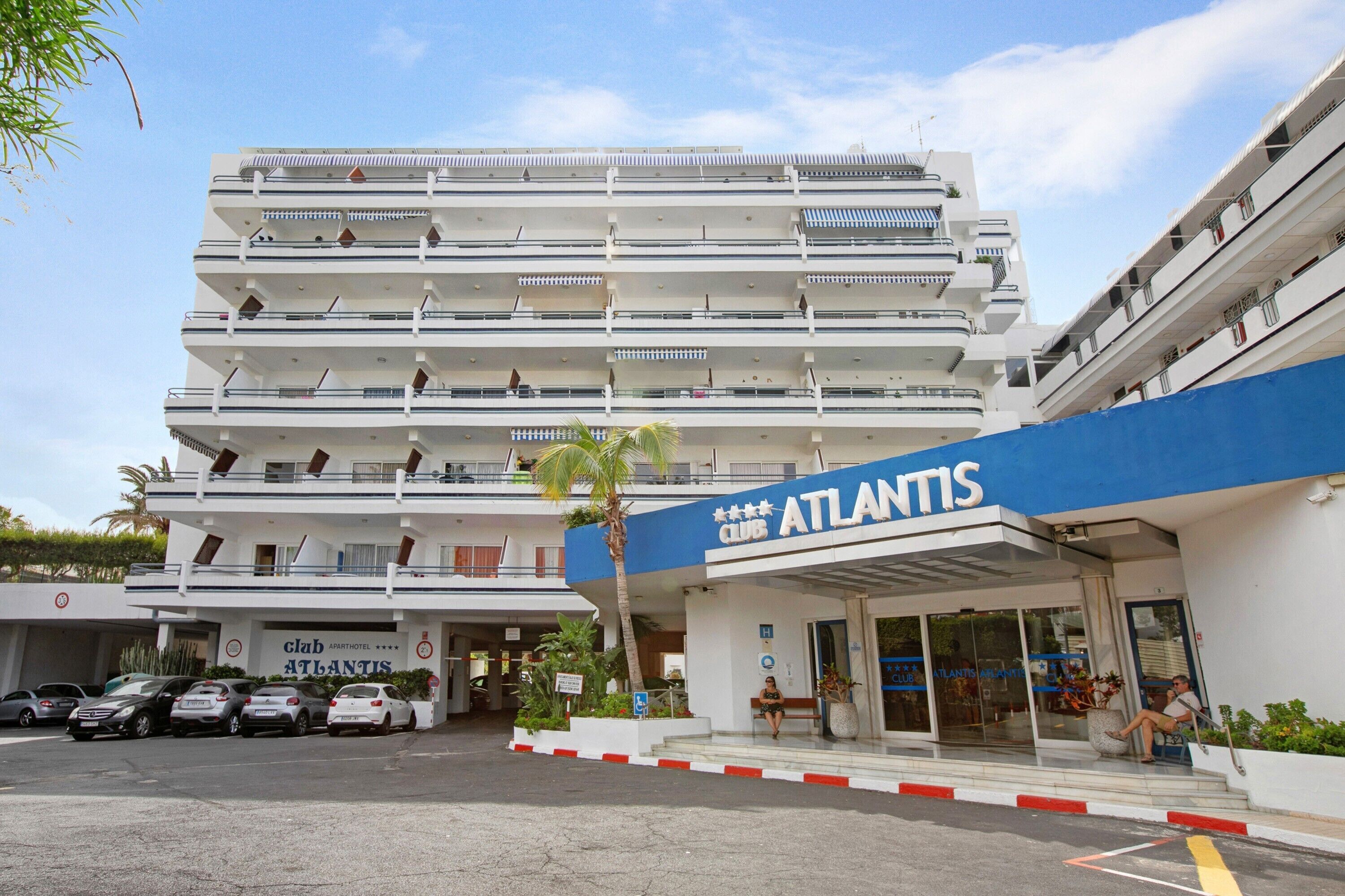 Un hôtel à plusieurs étages, appelé Atlantis, avec une enseigne bleue bien visible, un parking où sont garées plusieurs voitures, et un palmier au premier plan.
