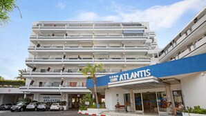 A multi-story hotel named Atlantis with a prominent blue sign, a parking area with several cars, and a palm tree in the foreground. - Vacation Home "Club Atlantis 223" with Sea View, Shared Pool & Wi-Fi (Adeje)