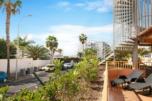 A balcony with lounge chairs, a street with cars, palm trees, and a building with a white facade. - Holiday Apartment 'Los Geranios 304' with Shared Pool, Private Terrace and Wi-Fi (Adeje)