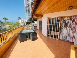 Un balcón con mesa y sillas, techo de madera y vista a palmeras y edificios.