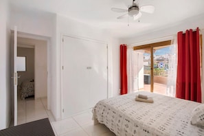 A bedroom with a bed, a ceiling fan, a balcony with red curtains, and a view of buildings. - Holiday Apartment 'Los Geranios 304' with Shared Pool, Private Terrace and Wi-Fi (Adeje)