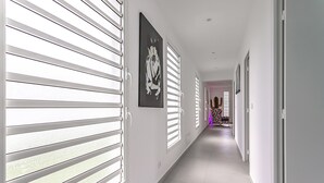 Un couloir aux murs blancs, doté d’un sol carrelé et de stores aux fenêtres. Un tableau encadré orne le mur, et une porte se trouve au fond du couloir.