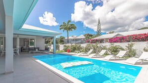 Une piscine avec des chaises longues, un espace de détente couvert et une vue sur les maisons voisines, ornées de fleurs roses.