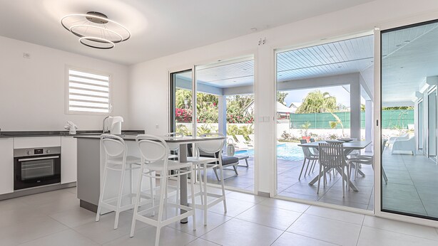Une cuisine moderne avec des meubles blancs et un four encastré, un coin repas avec des chaises et une table blanches, et une porte-fenêtre coulissante donnant sur une piscine.