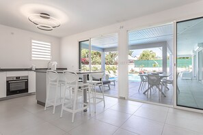 Una cocina moderna con armarios blancos y un horno empotrado, un comedor con sillas blancas y una mesa, y un vidrio corredizo que da a una zona de piscina.