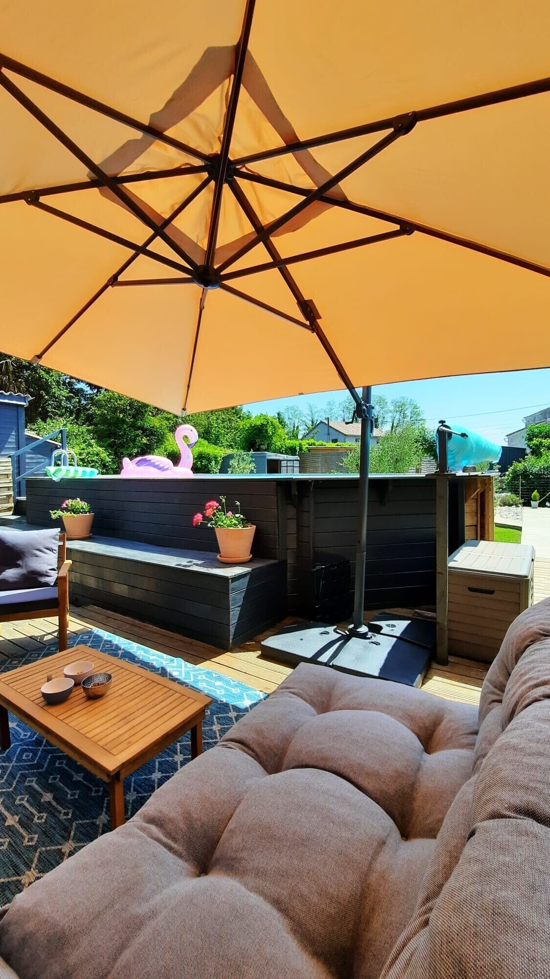 An outdoor patio with a large umbrella, a wooden table with bowls, a cushioned seating area, and a decorative flamingo.
