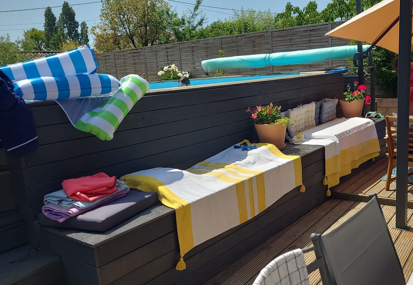Une terrasse en bois avec une piscine, un banc avec des serviettes et un espace couvert avec une table et des chaises.