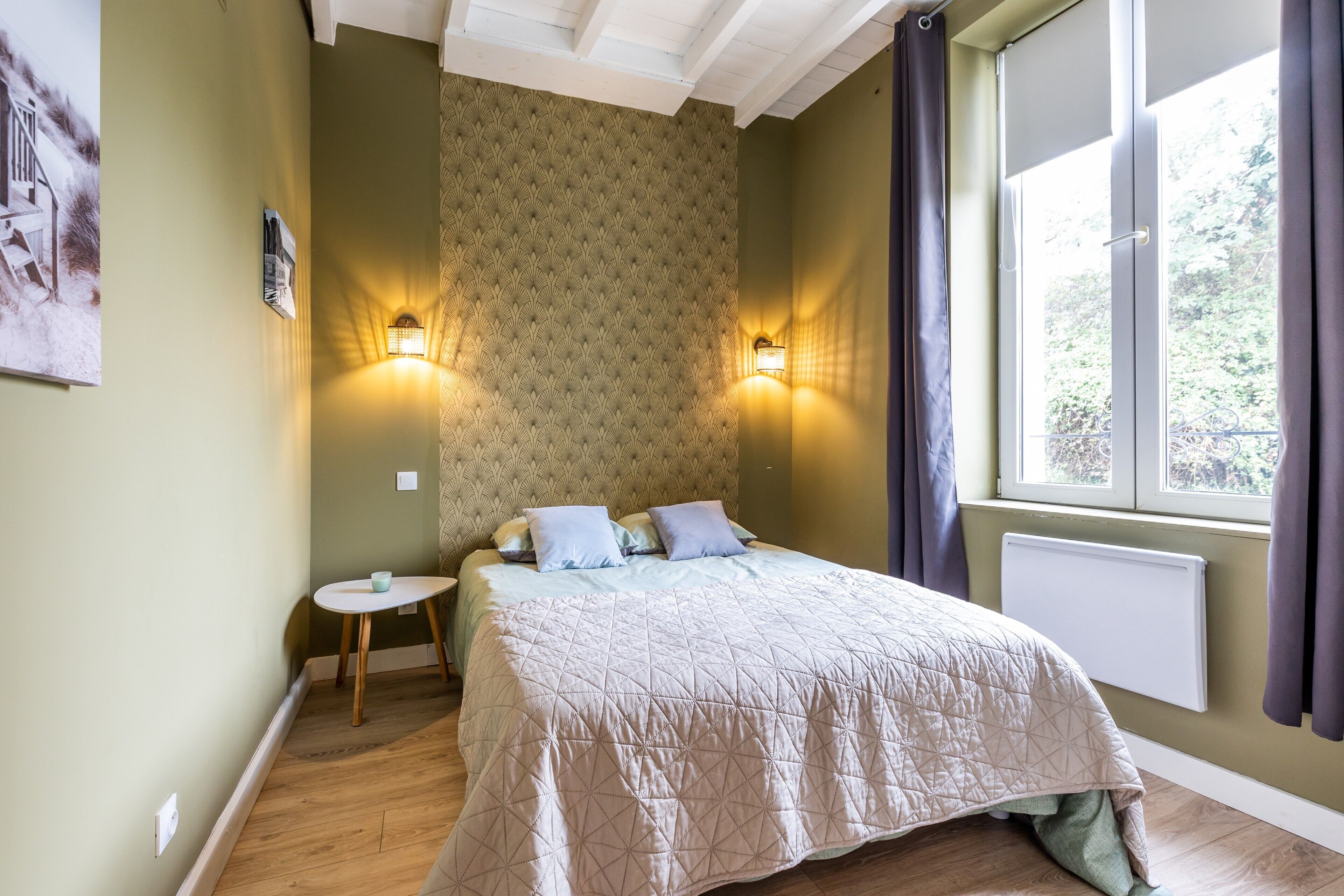 Un dormitorio con cama, lámparas de noche, una mesita y una ventana con cortinas.