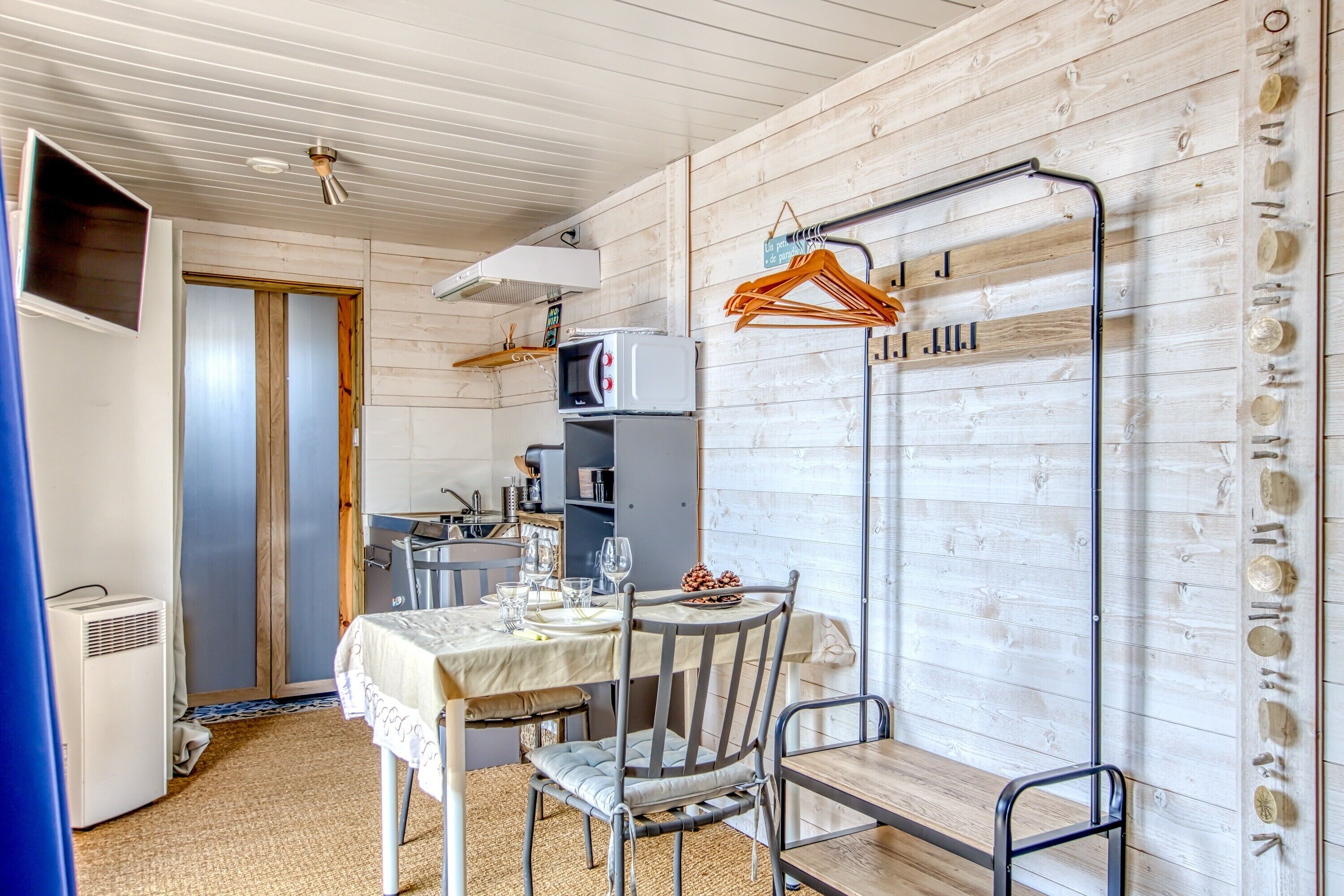 A room with a dining table set for two, a microwave, a small kitchen area, and a coat rack with hangers.