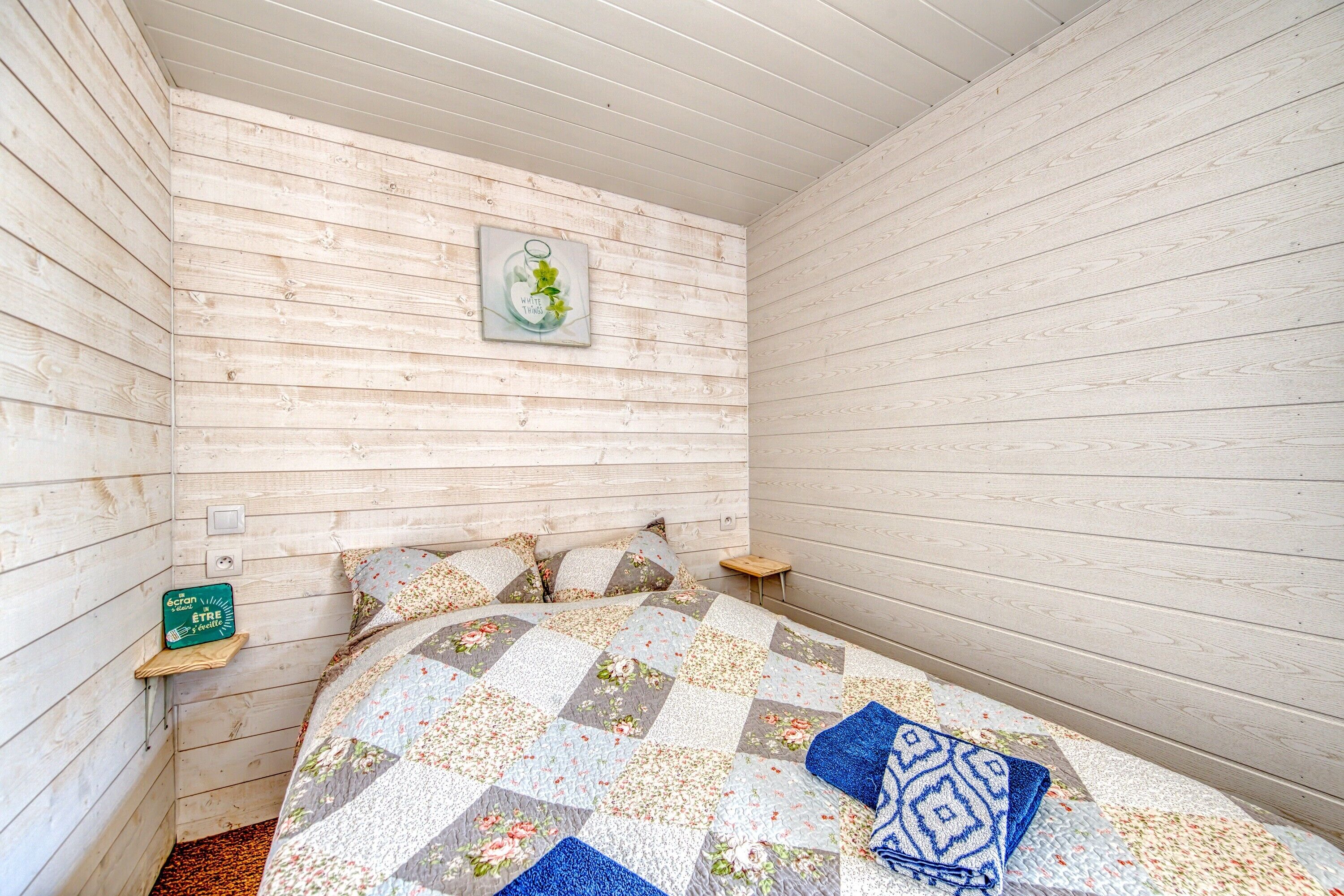 A bedroom with a wooden wall, a bed with a floral quilt, a small shelf with a sign, and a framed picture on the wall.