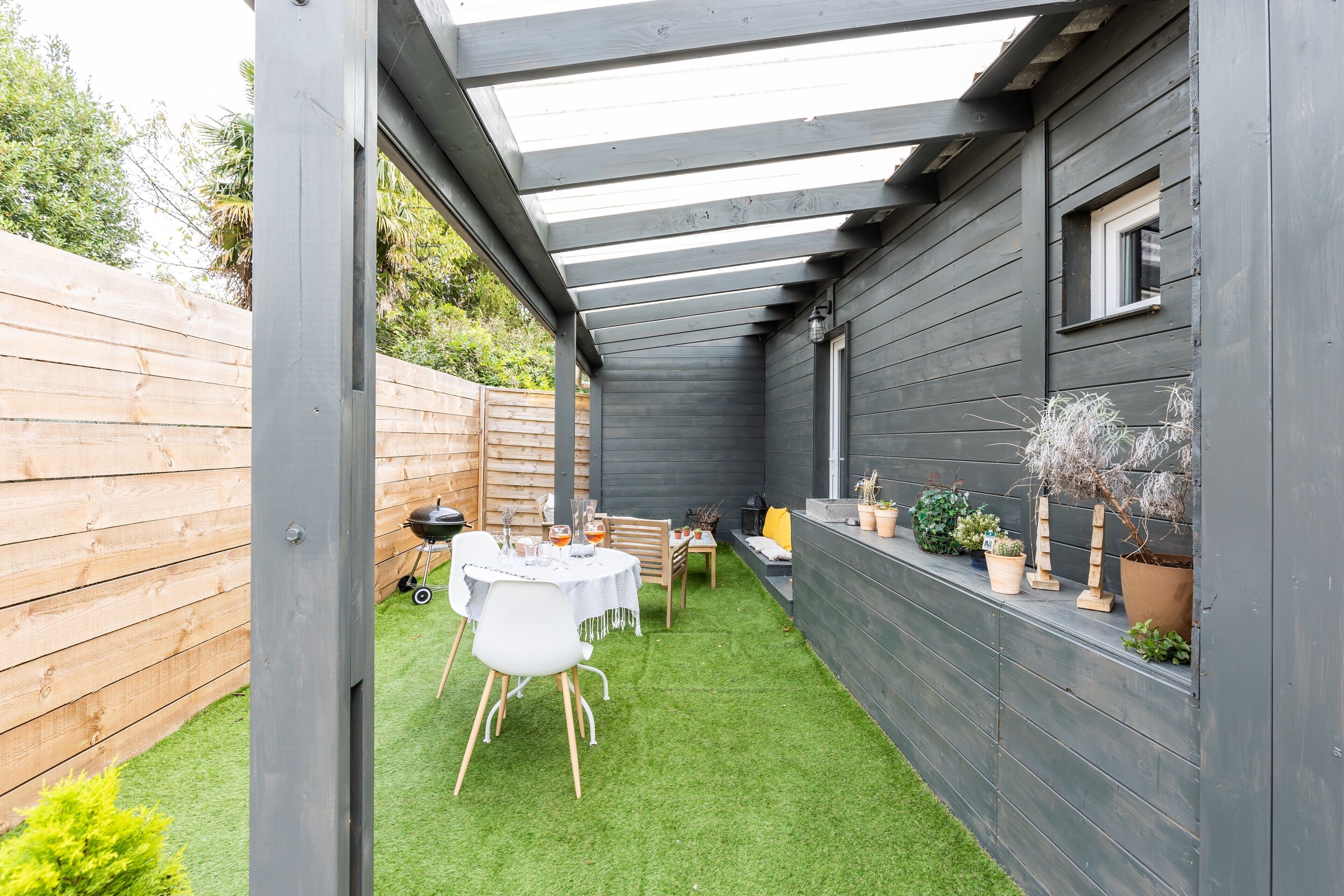A covered patio area with a table and chairs, a wooden fence, and potted plants.