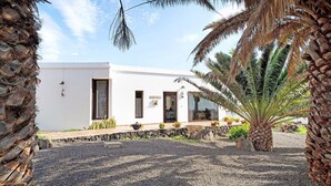 A white building with a gravel driveway, palm trees, and a stone wall. - Holiday Home 'Guenia I' with Mountain View, Wi-Fi and Air Conditioning (Teguise)
