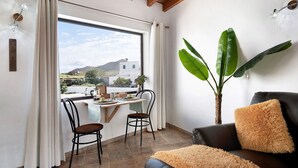 A room with a dining area, a black leather sofa, a large plant, and a view of buildings and mountains through a window. - Holiday Home 'Guenia I' with Mountain View, Wi-Fi and Air Conditioning (Teguise)