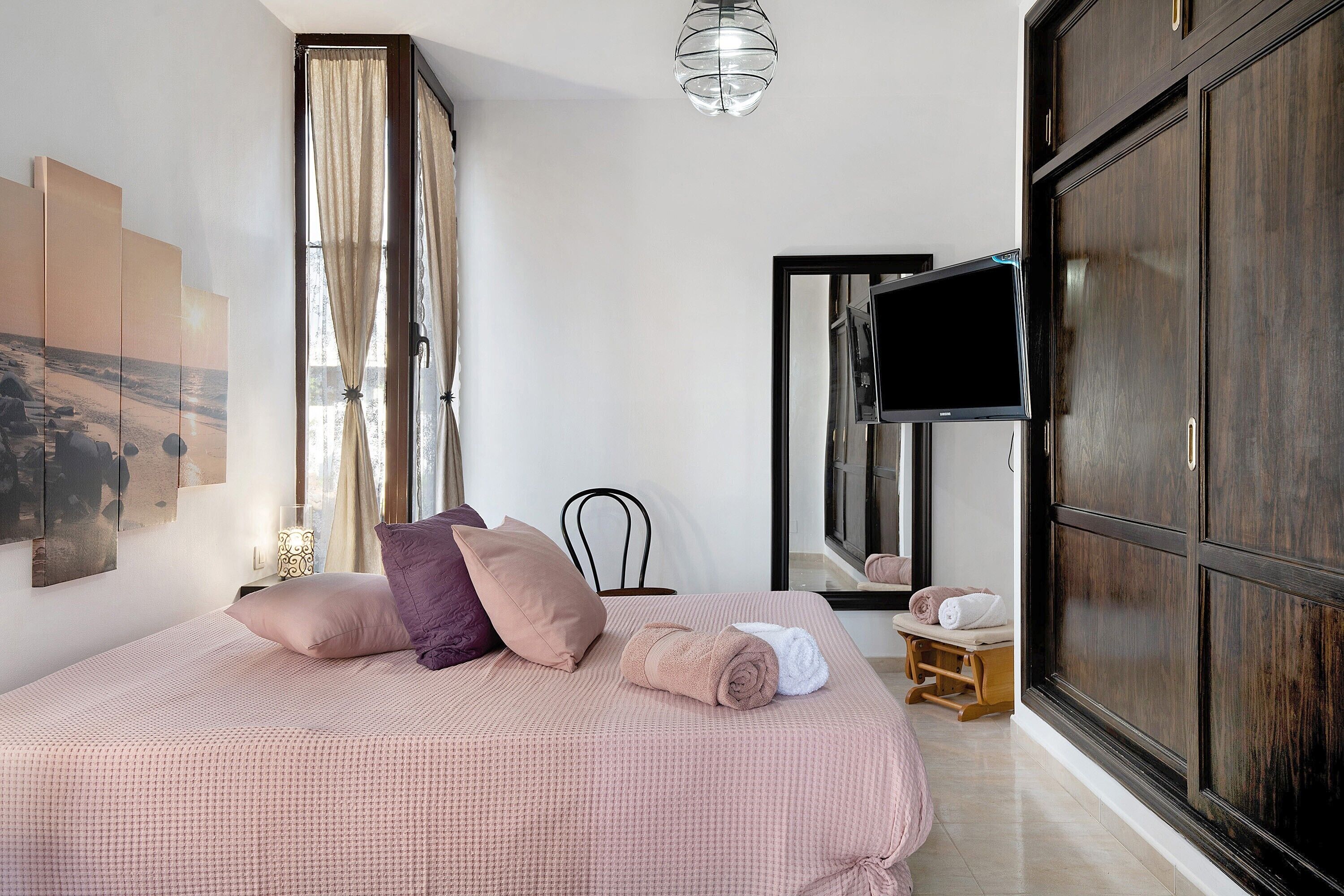 Une chambre à coucher avec un lit, des oreillers, une télévision, un miroir et une fenêtre avec des rideaux.