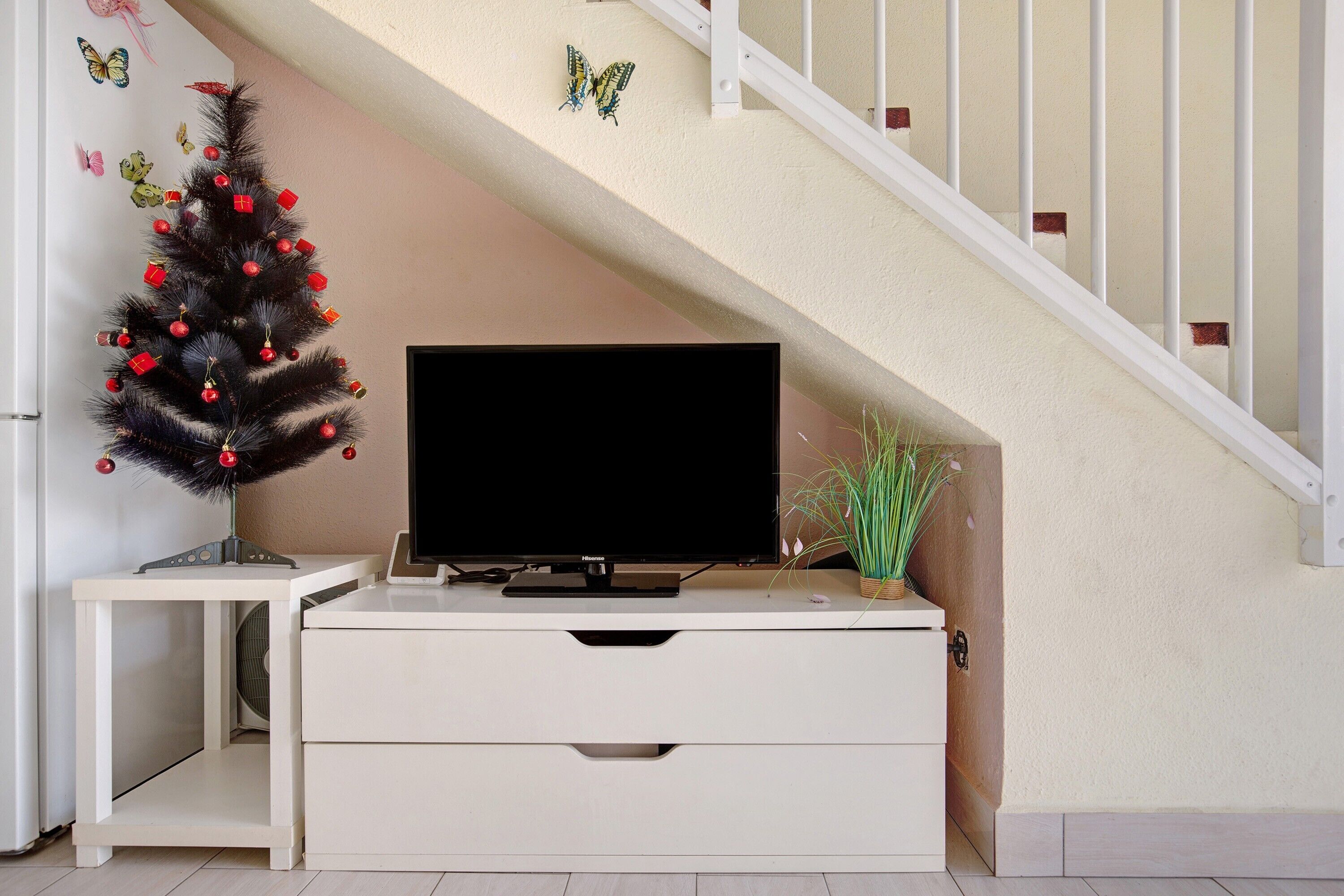 Eine Treppe mit weißer Geländer, ein kleiner Weihnachtsbaum mit Verzierungen, ein Fernseher auf einem weißen Schrank und eine Pflanze auf dem Boden.