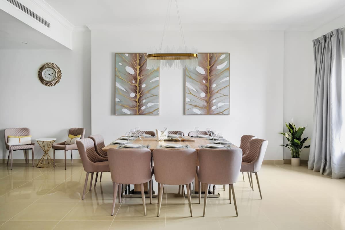Een eetkamer met een ronde tafel gedekt voor zes personen, omringd door zes stoelen. Een wandklok en een groot abstract schilderij hangen boven de tafel.