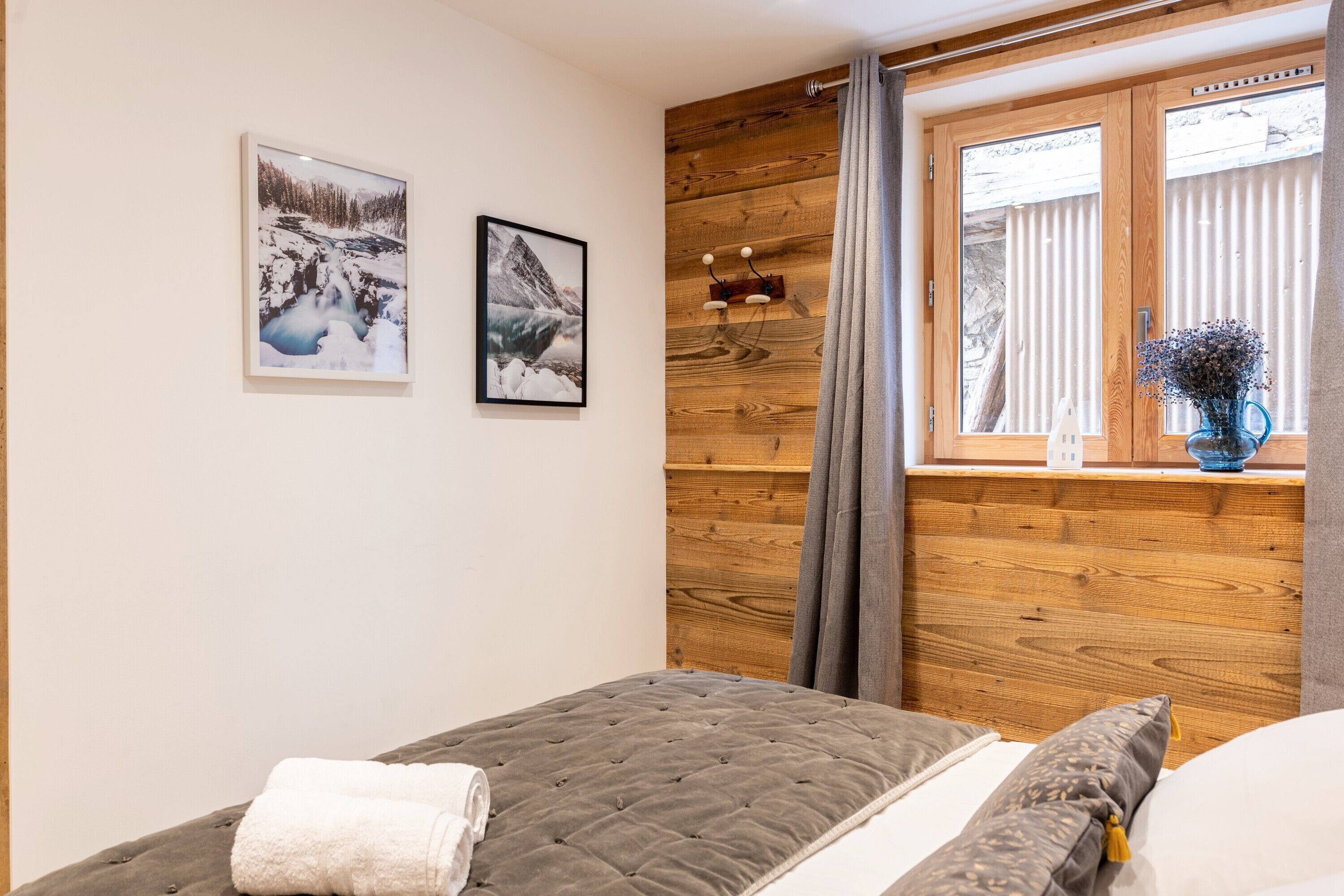 Une chambre avec une tête de lit en bois, une fenêtre avec des rideaux, un lit recouvert d’une couette grise et deux tableaux encadrés accrochés au mur.