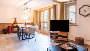 A modern living room with a flat-screen TV, wooden dining table, chairs, and a large window with curtains.