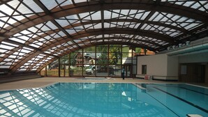 An indoor swimming pool with a glass ceiling, surrounded by a wooden deck and a view of houses.