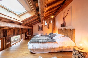 Un dormitorio con una cama grande, vigas de madera, un tragaluz, un cuadro de un ciervo y una mesa de troncos con una lámpara.