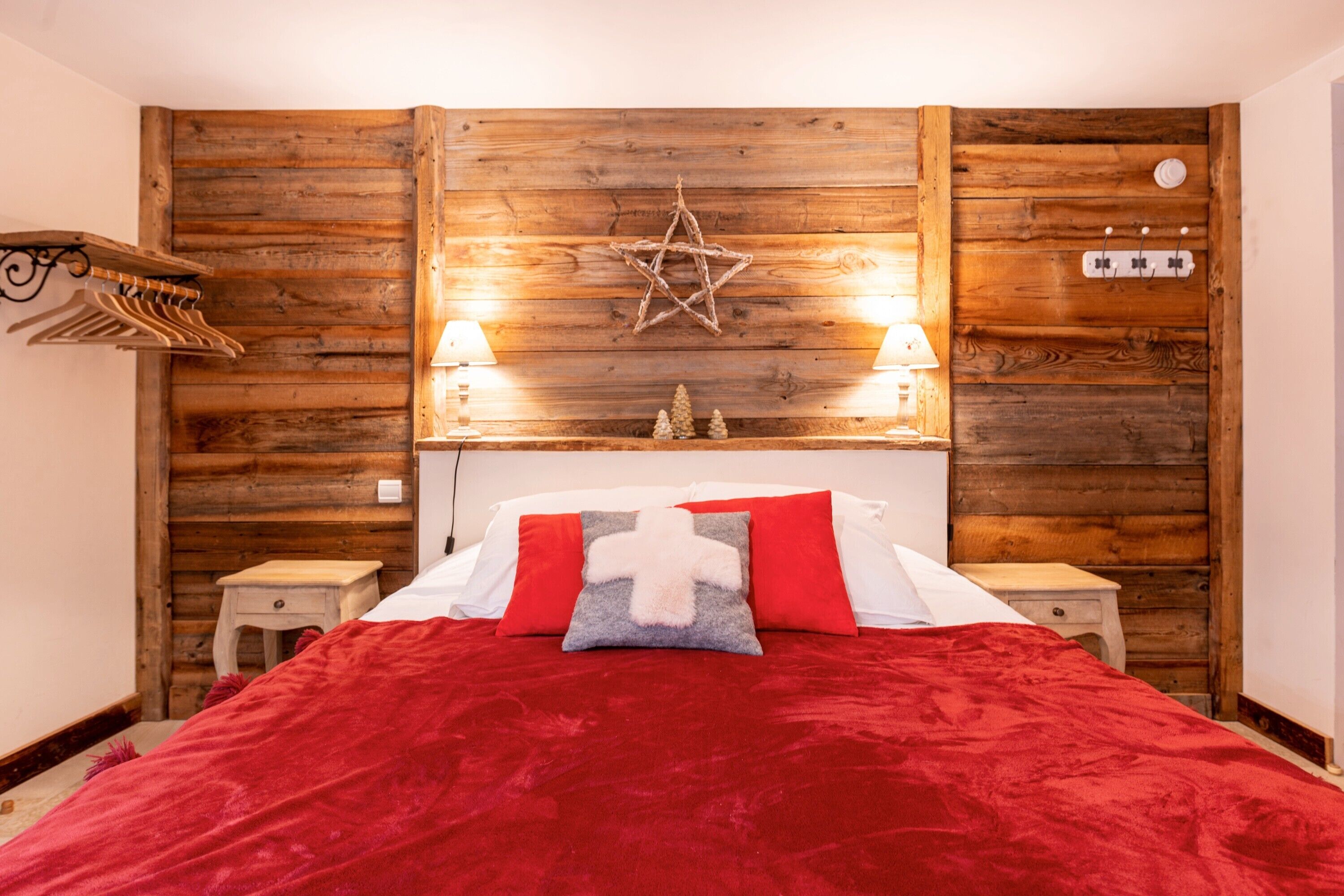 A room with a wooden wall, a bed with red bedding, two bedside lamps, a small table, and a wall-mounted star decoration.