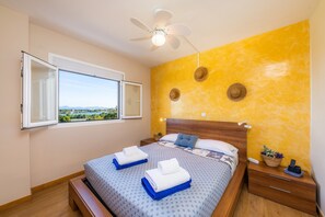 Un dormitorio con una cama, una ventana con cortina abierta, un ventilador de techo y una mesita de noche con una cesta.