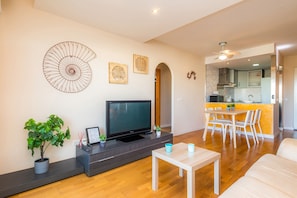 A modern living room with a flat-screen TV, a dining area, and a kitchen visible through an archway.