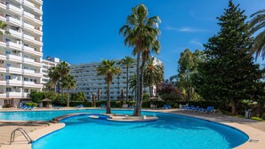 A swimming pool with palm trees, lounge chairs, and a multi-story building in the background. - Apartment "Sweet Sea" in Port of Alcudia with Shared Pool, Shared Terraces & Wi-Fi (Puerto de Alcudia)