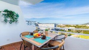 A balcony with a table set for breakfast, wicker chairs, and a view of buildings and greenery. - Apartment "Sweet Sea" in Port of Alcudia with Shared Pool, Shared Terraces & Wi-Fi (Puerto de Alcudia)