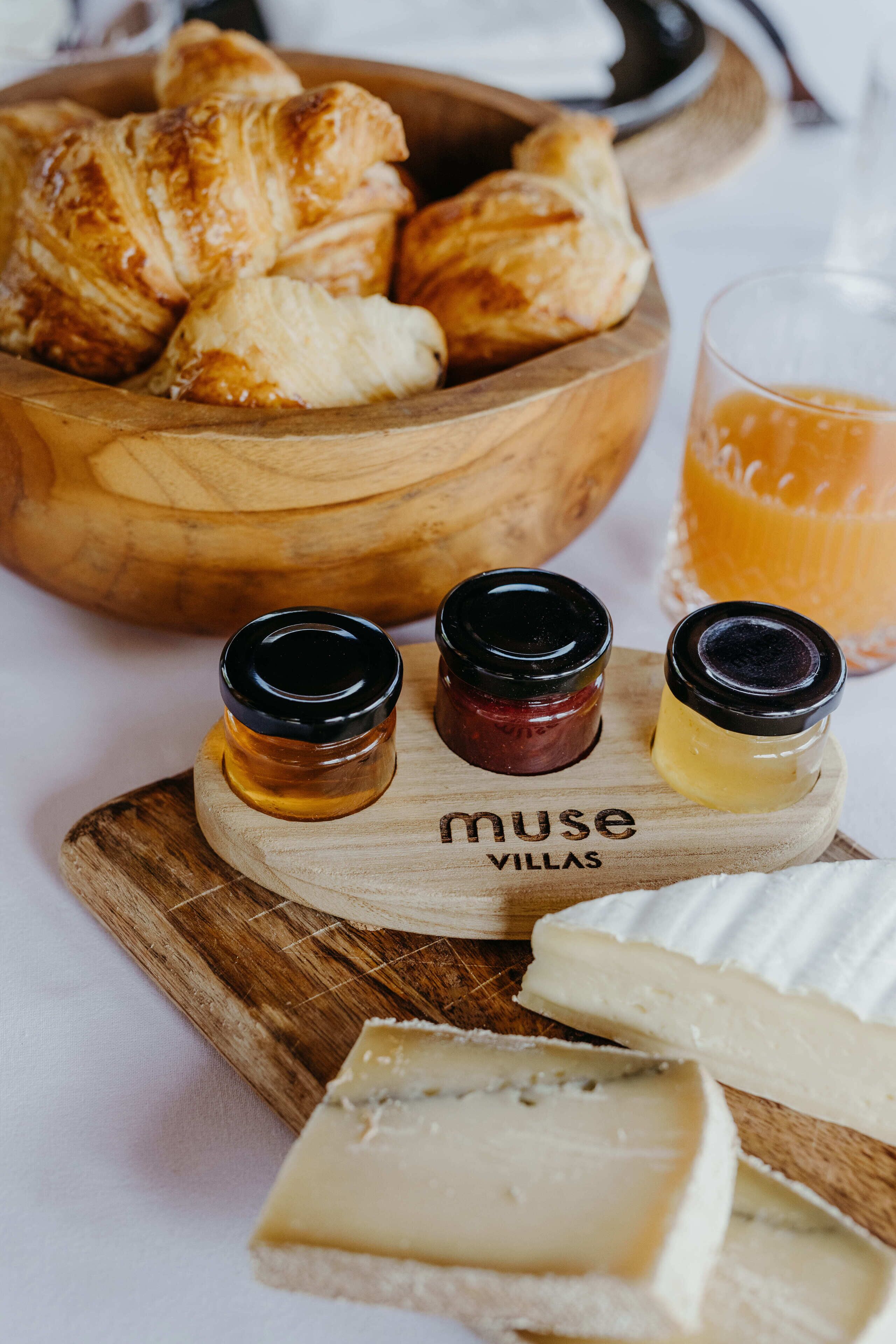 A wooden board with cheese, jars of jam, and croissants.