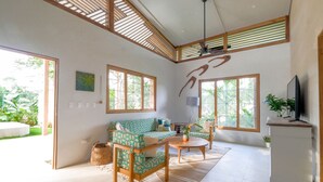 Een ruime woonkamer met een bank, salontafel en een televisie. Grote ramen zorgen voor veel natuurlijk licht.