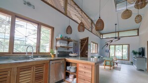 Een ruime keuken met houten kastjes, een ingebouwde vaatwasser en een groot kookeiland met een spoelbak. Er zijn rieten hanglampen, een groot raam met uitzicht, en een eethoek met een tafel en stoelen.