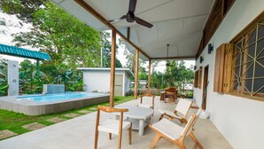 Een overdekt terras met een zwembad, houten stoelen en een plafondventilator.