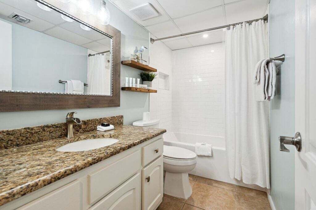 Ein Badezimmer mit Granitwaschtisch, Toilette in Weiß, Dusche mit weißem Vorhang und Spiegel mit Holzrahmen.