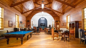 Une pièce spacieuse aux murs et au plafond en bois, avec une table de billard, plusieurs coins salon et une grande fenêtre en arc de cercle.