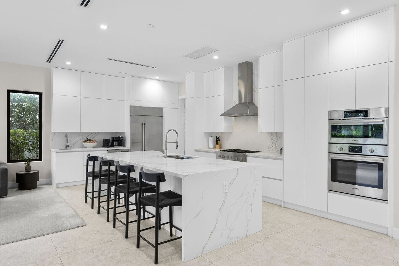 Une cuisine moderne avec des meubles blancs, un îlot central doté de tabourets, un four encastré et une grande fenêtre.