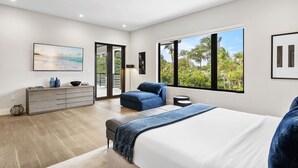 A bedroom with a large bed, a seating area, a dresser, and a view of greenery through the windows.