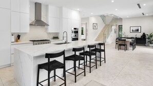 A modern kitchen with white cabinetry, a marble island, black bar stools, a staircase, and a dining area with a black table and chairs.