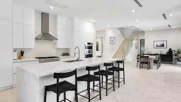 Une cuisine moderne avec des meubles blancs, un îlot en marbre, des tabourets de bar noirs, un escalier et un coin repas avec une table et des chaises noires.