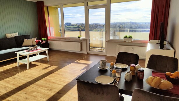 Ein modernes Wohnzimmer mit Essbereich, großen Fenstern und Blick auf eine entfernte Landschaft, einer Couch, einem Couchtisch und einem für ein Essen gedeckten Esstisch.