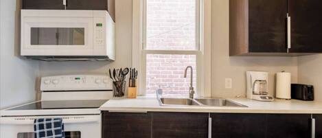 A kitchen with a white microwave, stove, and sink. There is a window with a brick wall view.