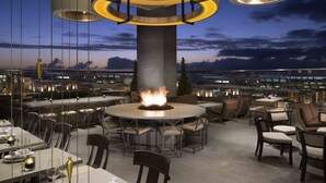 A rooftop dining area with a central fire feature, surrounded by tables and chairs, overlooking a cityscape at dusk.