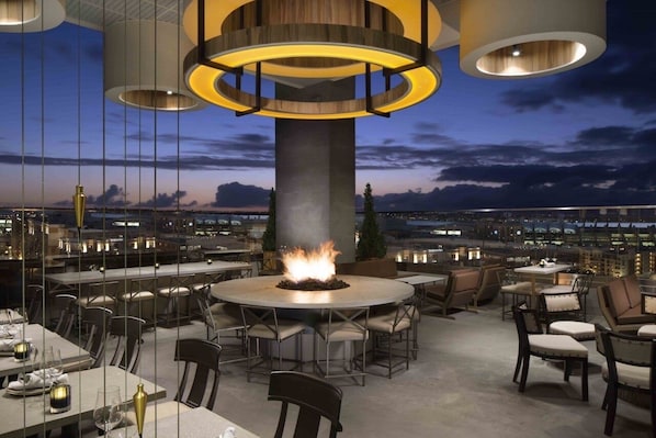 A rooftop dining area with a central fire feature, surrounded by tables and chairs, overlooking a cityscape at dusk.