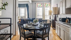 A modern kitchen with white cabinets, a dining table with chairs, and a living area with a sofa and blue curtains.