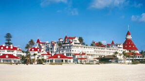 A beachfront hotel with multiple red-roofed buildings, a prominent red tower, and palm trees.
